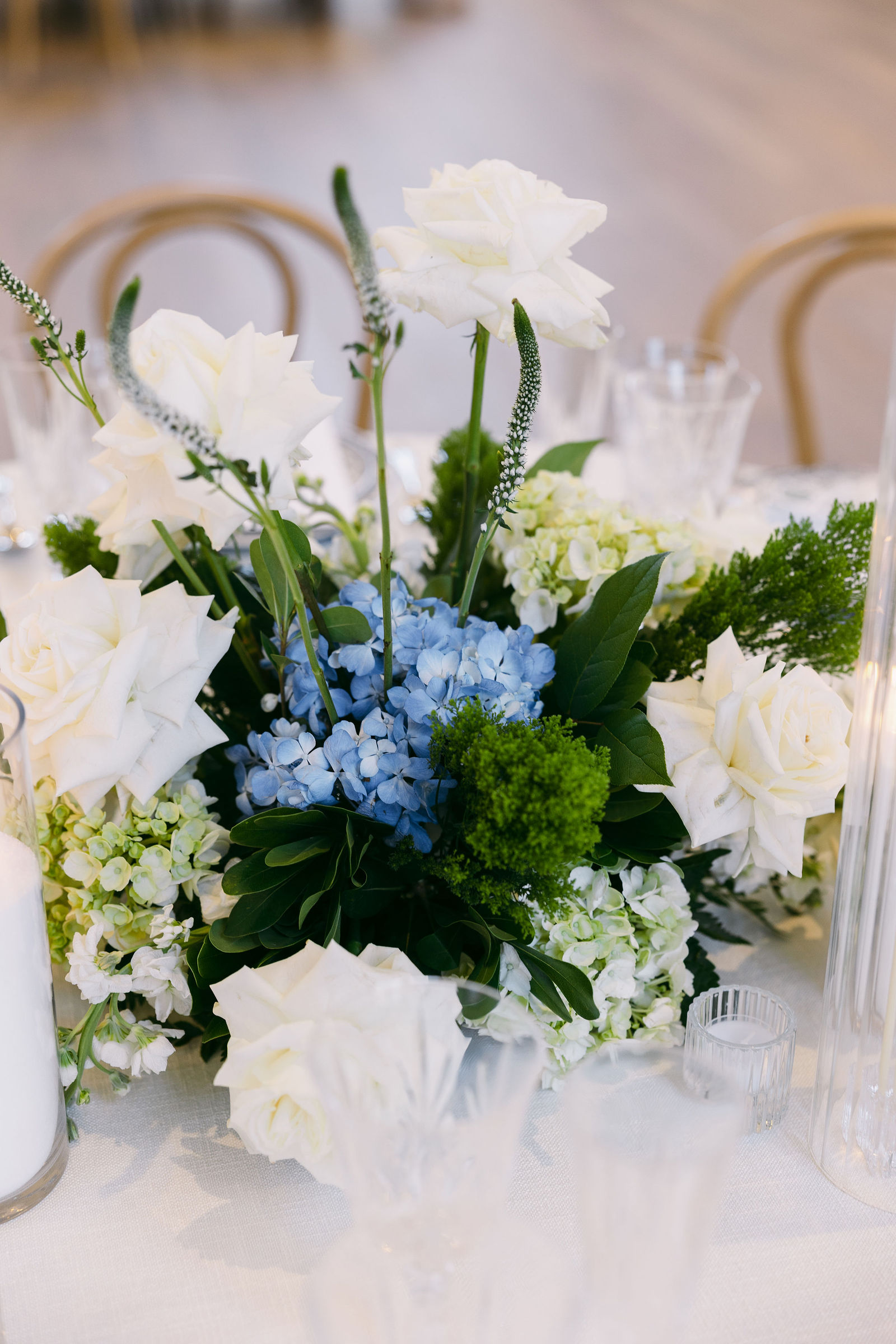Close-up of a centerpiece of white garden roses, blue hydrangeas, and lush greenery on a reception table